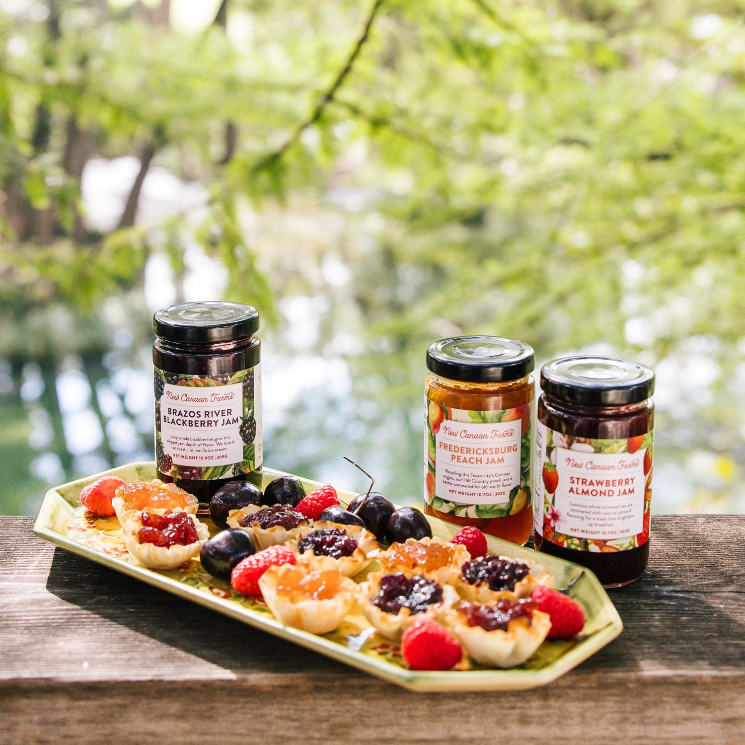 Three jars of jam on a tray with small pastries and berries outdoors.