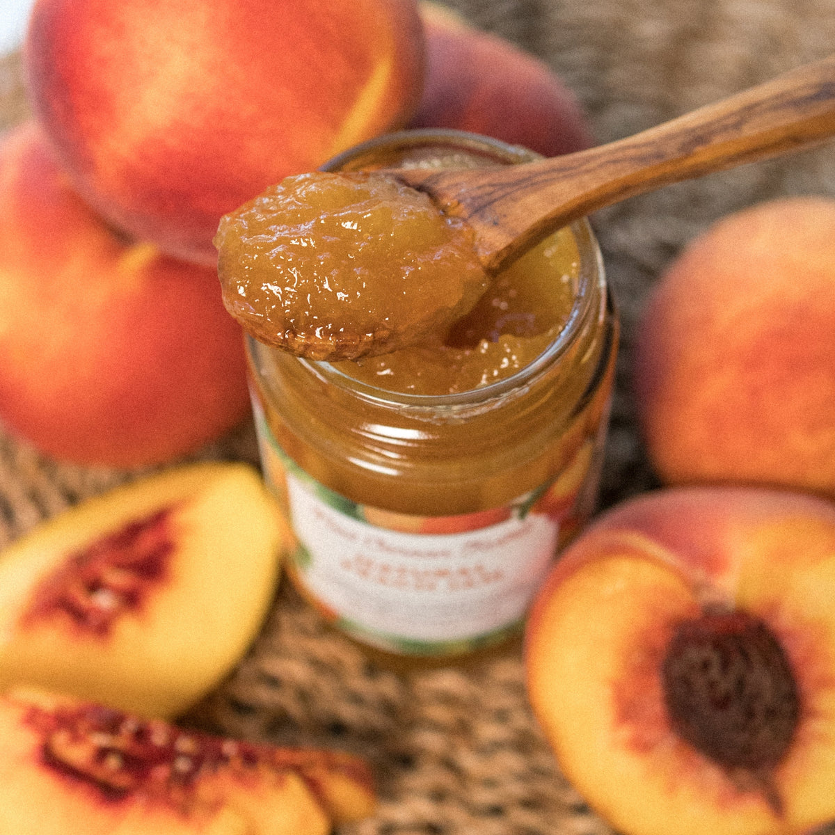 A wooden spoon of Natural Peach Jam over an open jar, with whole and sliced peaches on a woven mat