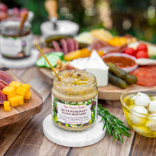 Jar of olive rosemary dijon mustard on a wooden table with various food items in the background