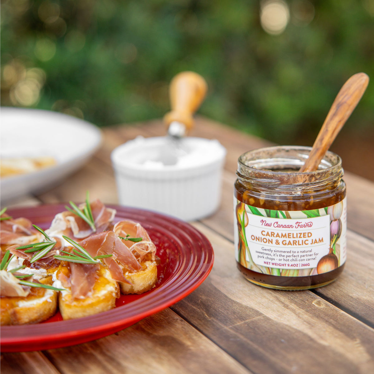 Jar of caramelized onion and garlic jam on a wooden table with crostini and rosemary.