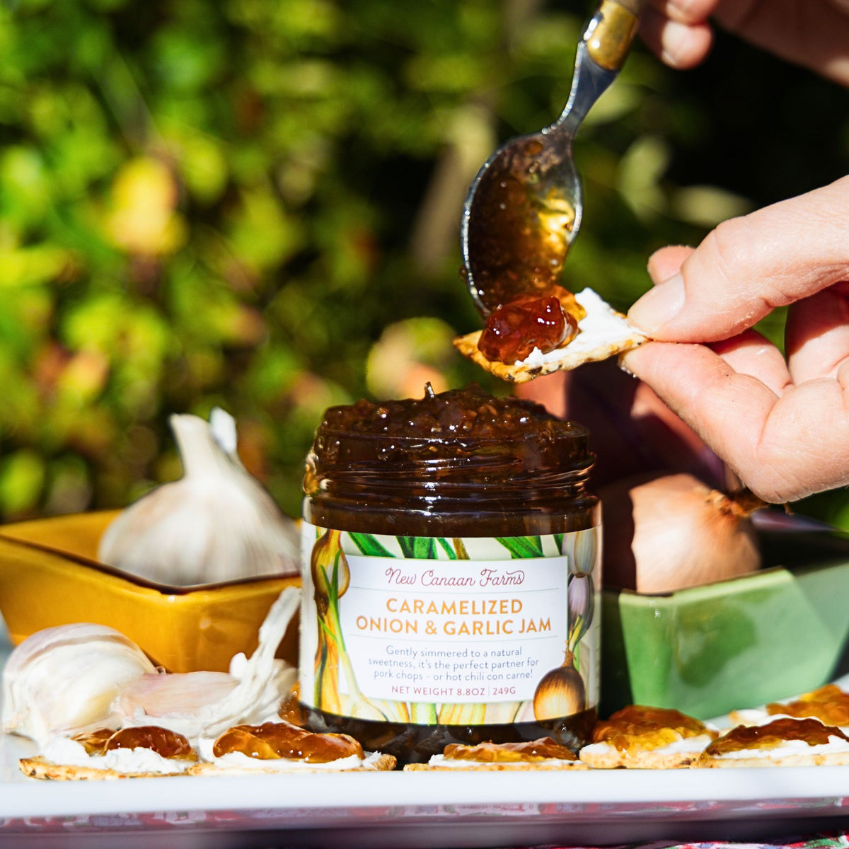 Person spreading caramelized onion and garlic jam on a cracker with a spoon, jar of jam in the foreground.