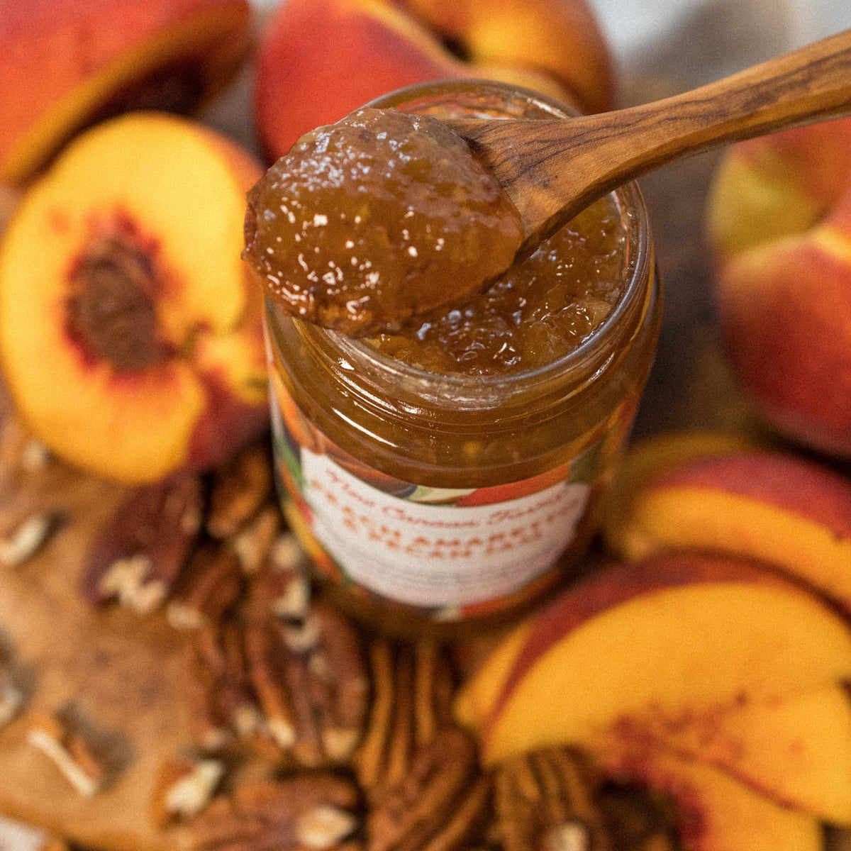 Close-up of a wooden spoon of Peach Amaretto & Pecan Jam over the open jar, with peaches and pecans
