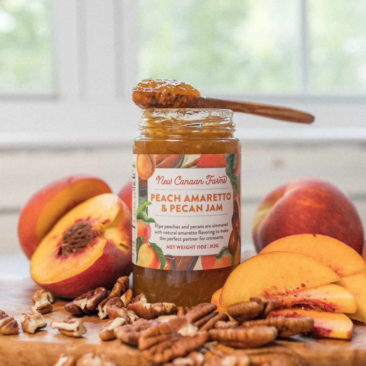 Jar of New Canaan Farms Peach Amaretto & Pecan Jam with a spoonful on top, among fresh peaches and pecans