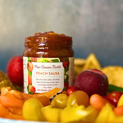 Jar of New Canaan Farms Peach Salsa surrounded by peaches and salsa on a blurred background