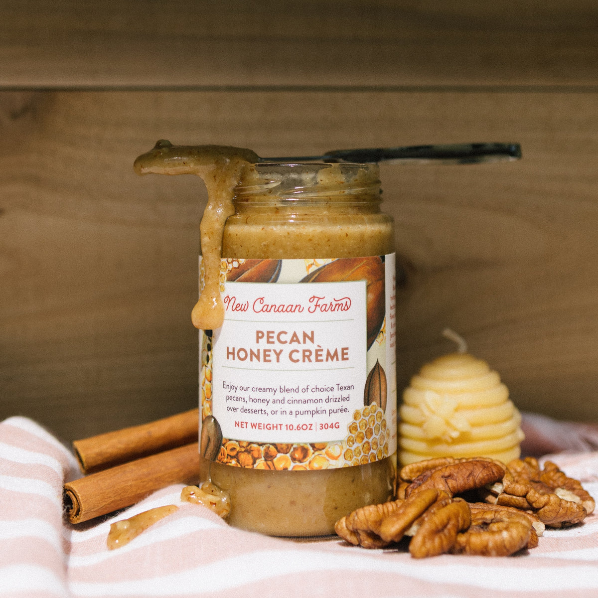 New Canaan Farms Pecan Honey Crème jar with spread dripping off a knife, beside cinnamon, pecans and a candle