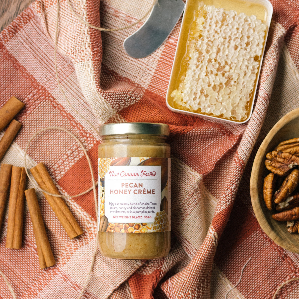 New Canaan Farms Pecan Honey Crème jar on a plaid cloth with cinnamon sticks, honeycomb and pecans