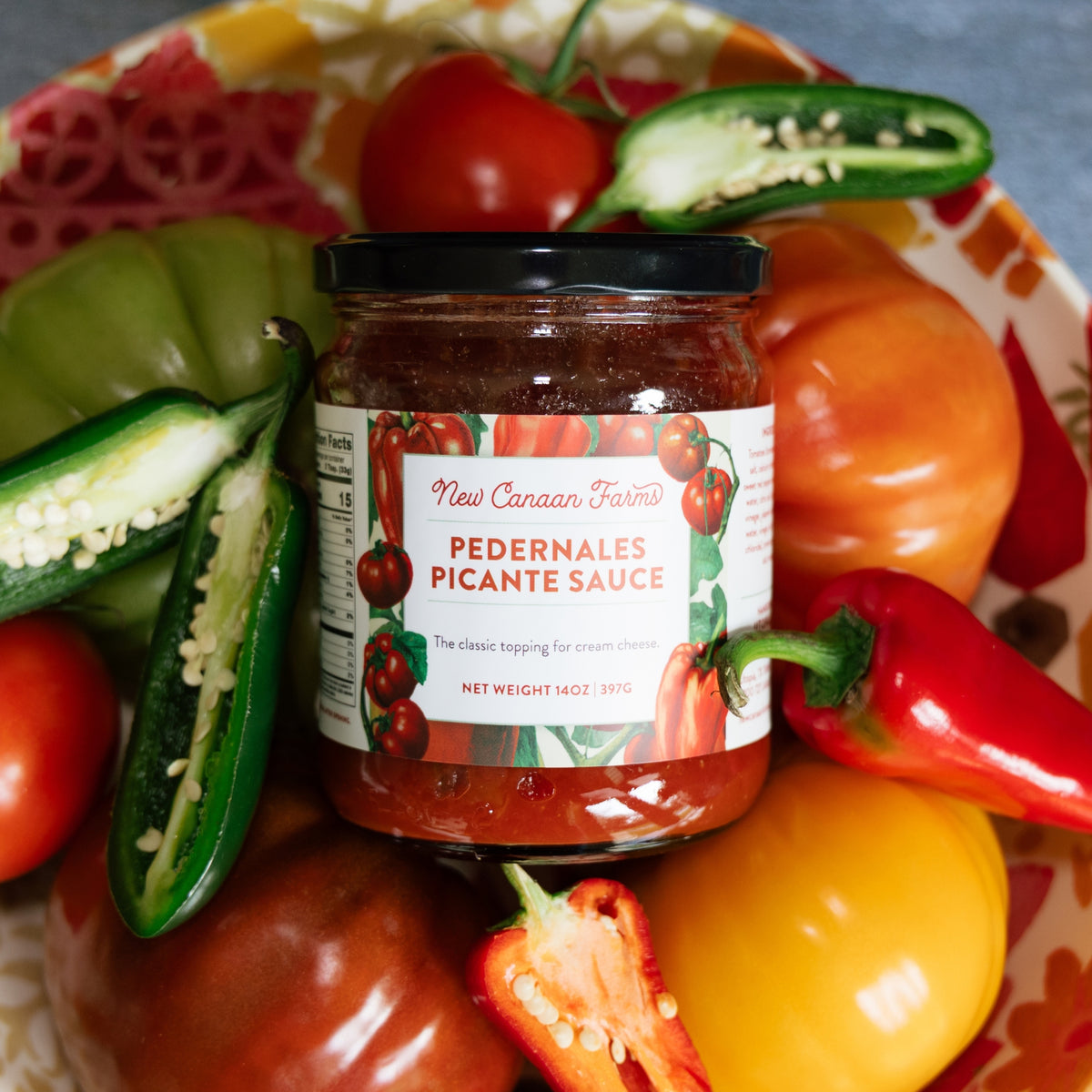 Jar of Pedernales Picante Sauce surrounded by peppers and tomatoes on a colorful plate.