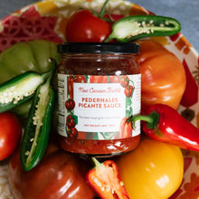 Jar of Pedernales Picante Sauce surrounded by peppers and tomatoes on a colorful plate.