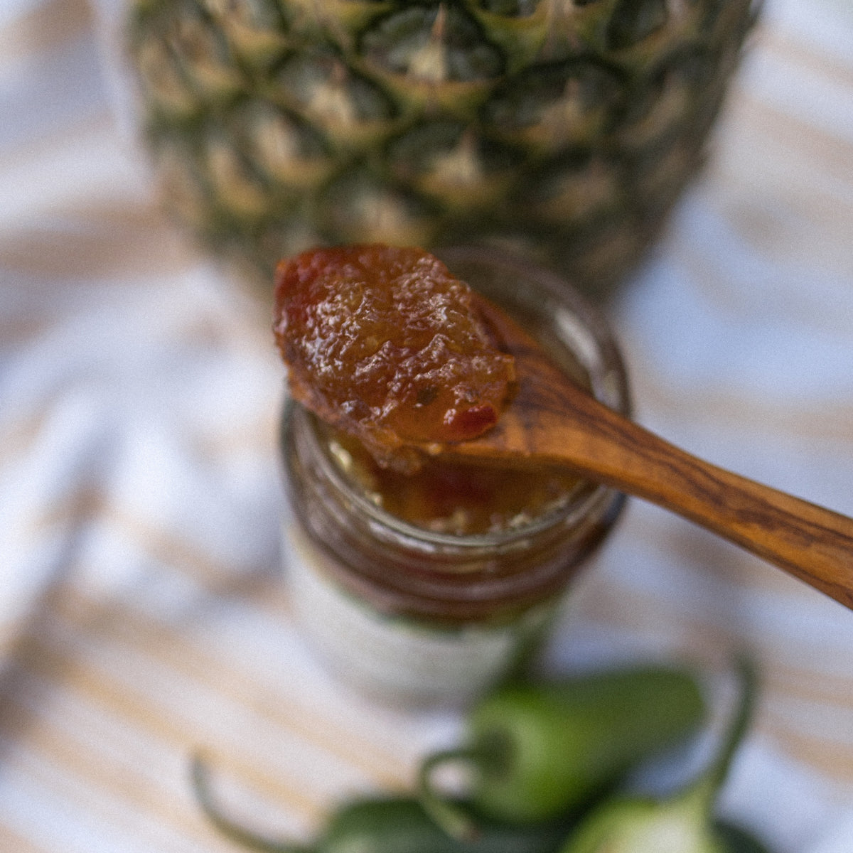 A wooden spoon of Pineapple Jalapeño Jelly over an open jar, with a pineapple and jalapeños behind