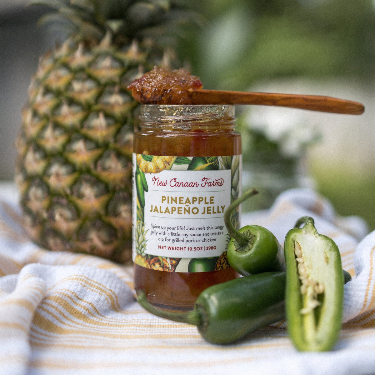 New Canaan Farms Pineapple Jalapeño Jelly jar with a spoonful on top, beside a pineapple and jalapeños