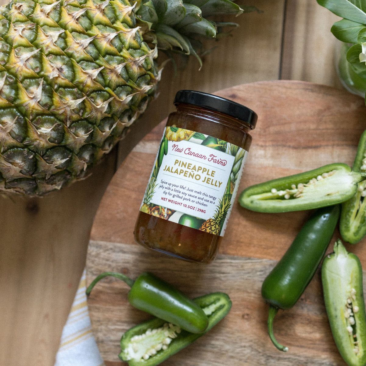 New Canaan Farms Pineapple Jalapeño Jelly jar on a wooden board with a pineapple and sliced jalapeños