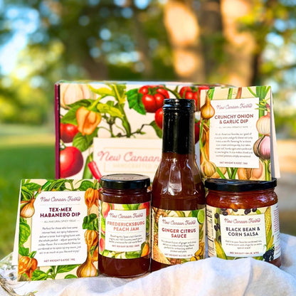 A curated collection of five New Canaan Farms  jams, sauces, salsas and spice blends, in front of a vegetable-designed gift box, photographed outside