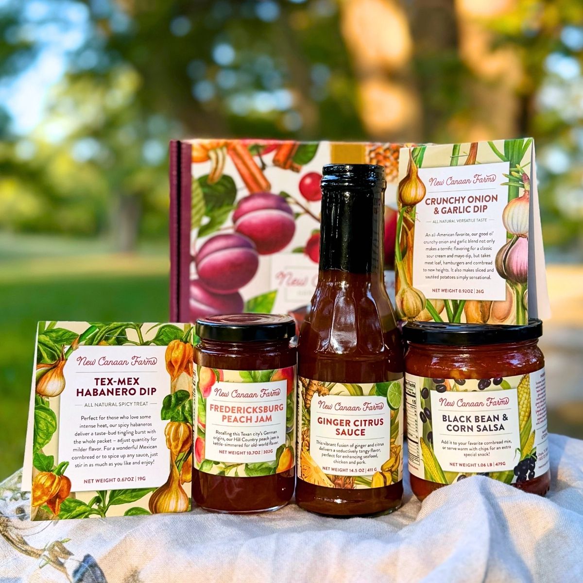 A curated collection of five New Canaan Farms  jams, sauces, salsas and spice blends, in front of a seasonally-inspired gift box, outside