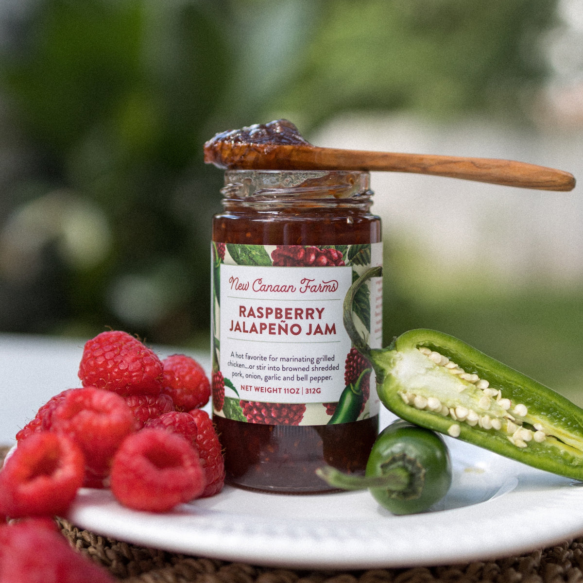 New Canaan Farms Raspberry Jalapeño Jam jar with a spoonful on top, beside raspberries and a halved jalapeño