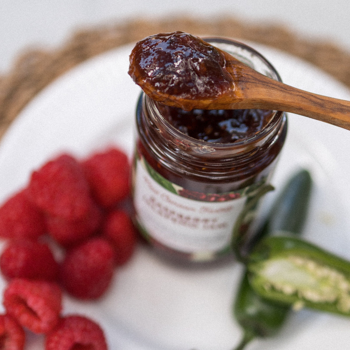 A wooden spoon of Raspberry Jalapeño Jam over an open jar, with raspberries and a halved jalapeño