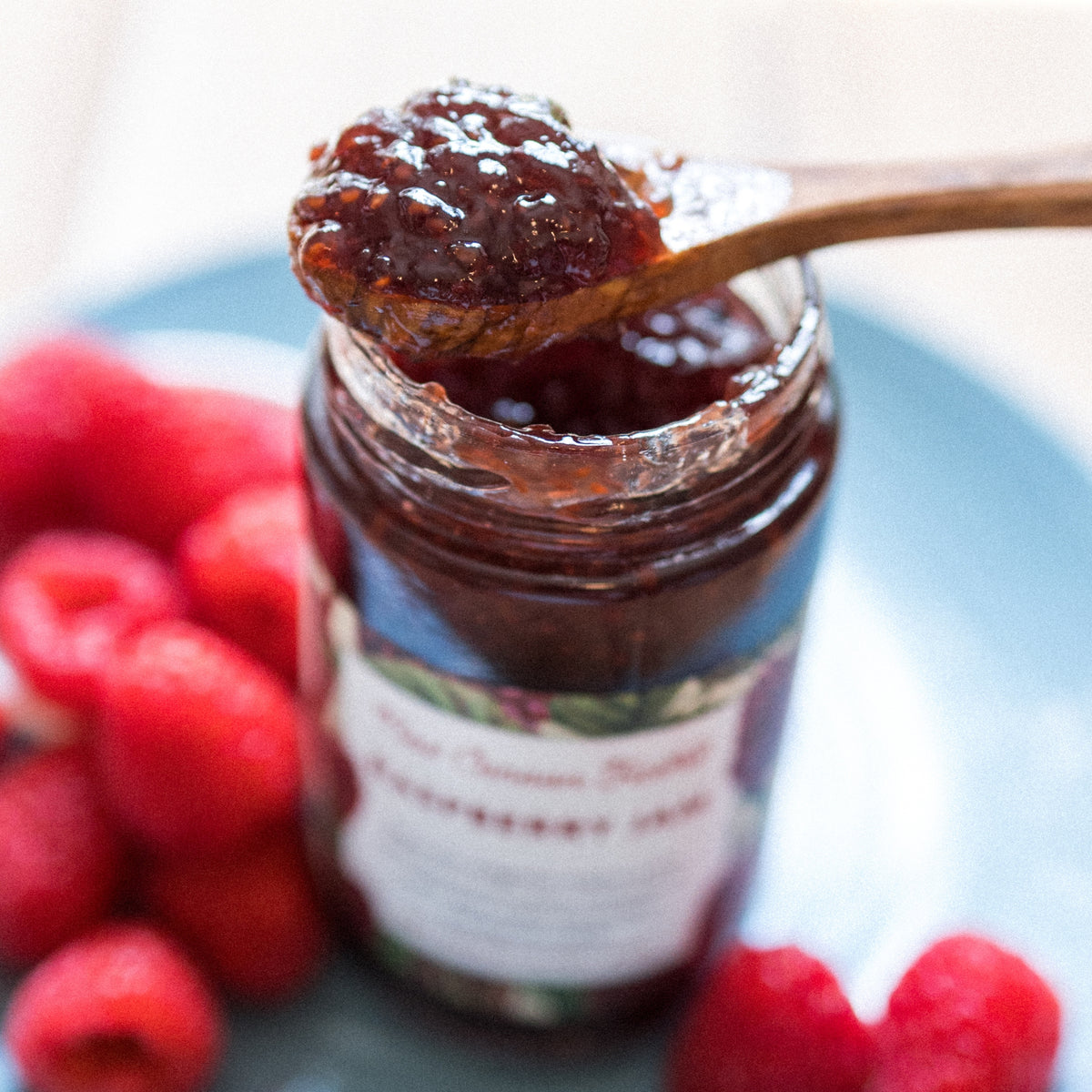 A jar of New Canaan Farms Raspberry Jam, with raspberries on a blue plate