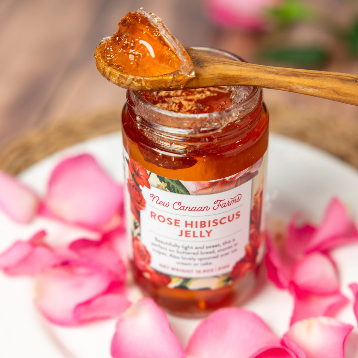 Jar of New Canaan Farms Rose Hibiscus Jelly with a spoonful of jelly on top, surrounded by pink petals.