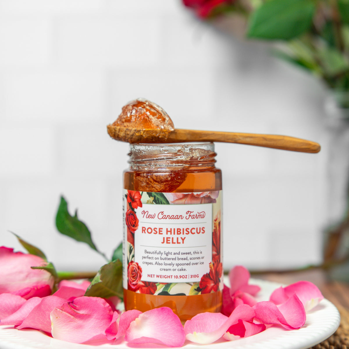 Jar of New Canaan Farms Rose Hibiscus Jelly with a wooden spoon on pink rose petals.
