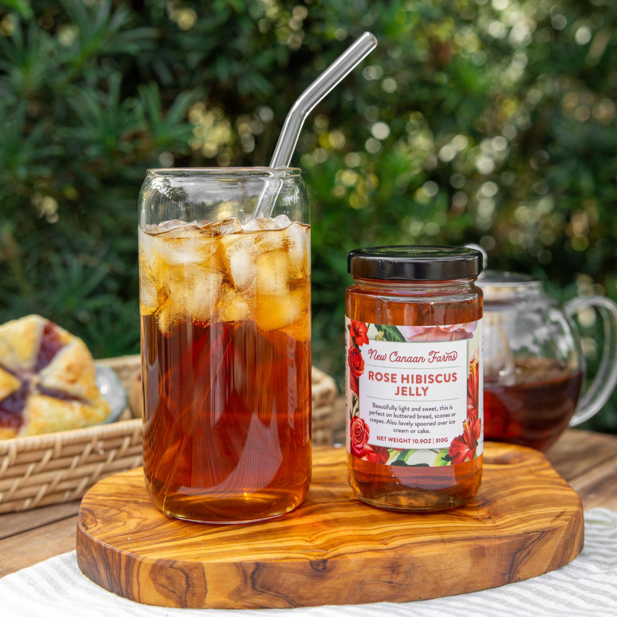 Glass of iced tea with a straw, jar of rose hibiscus jelly, and pastries on a wooden board outdoors.