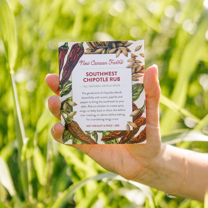 Hand holding a New Canaan Farms Southwest Chipotle Rub packet against a green outdoor background