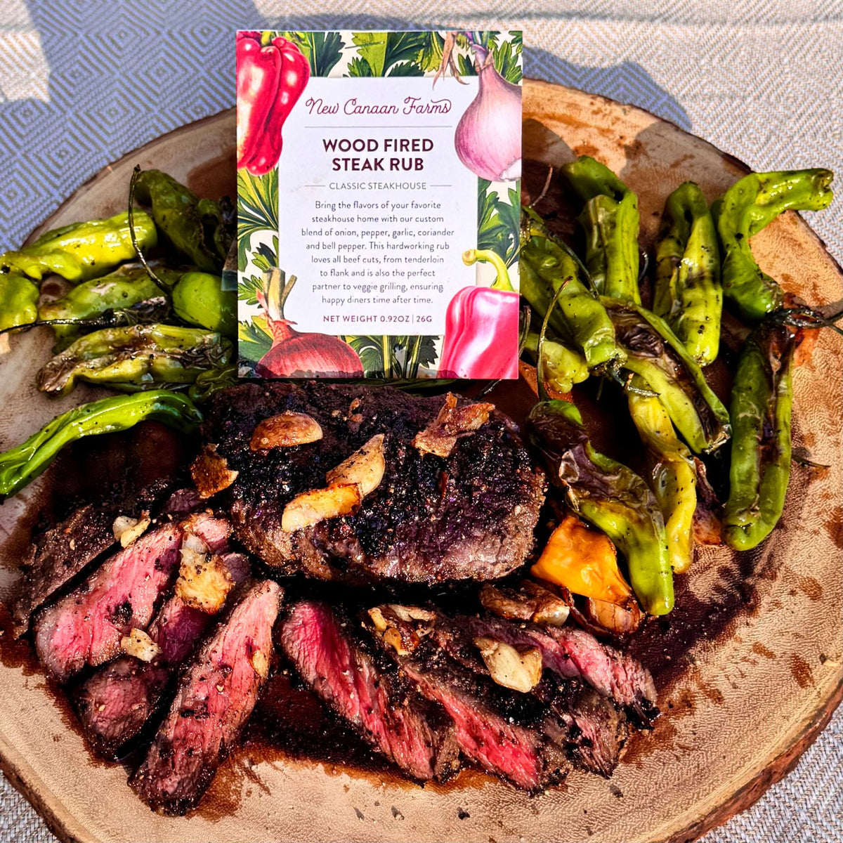 Wood-Fired Steak Rub packet propped above sliced seared steak with garlic chips and blistered green peppers on a wood board