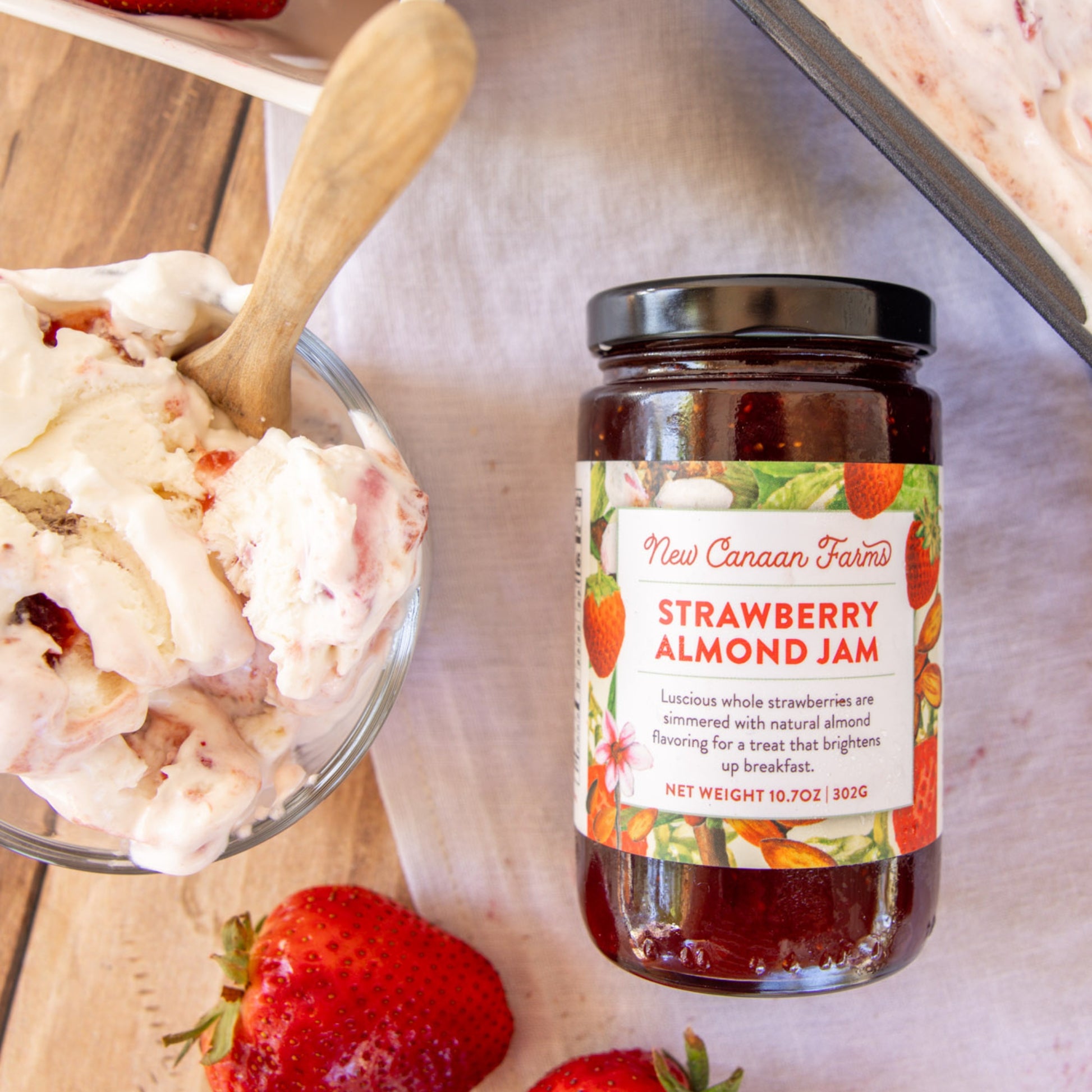 Jar of New Canaan Farms Strawberry Almond Jam with a bowl of ice cream and strawberries on a wooden surface.