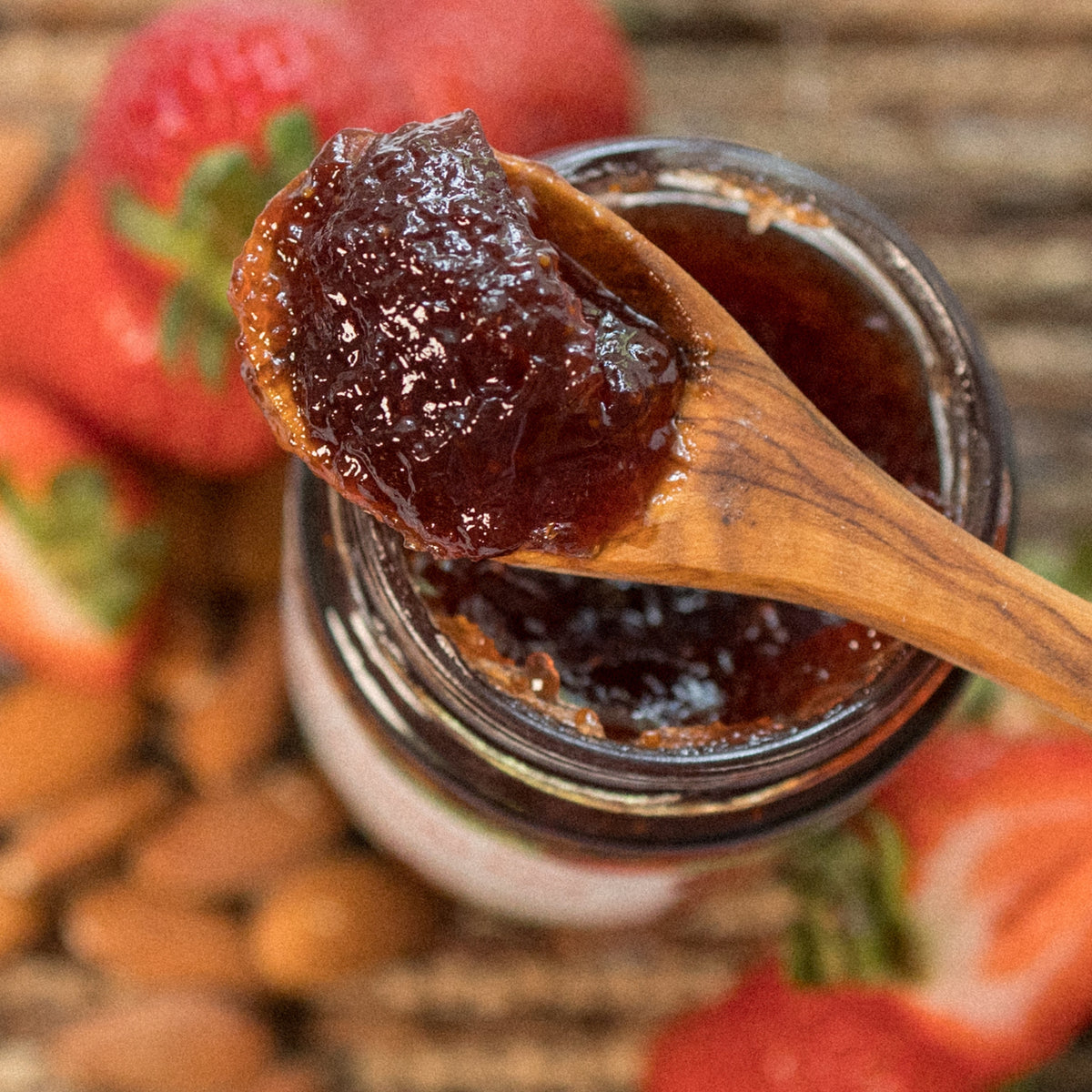 A wooden spoon of Strawberry Almond Jam over an open jar, with strawberries and almonds behind