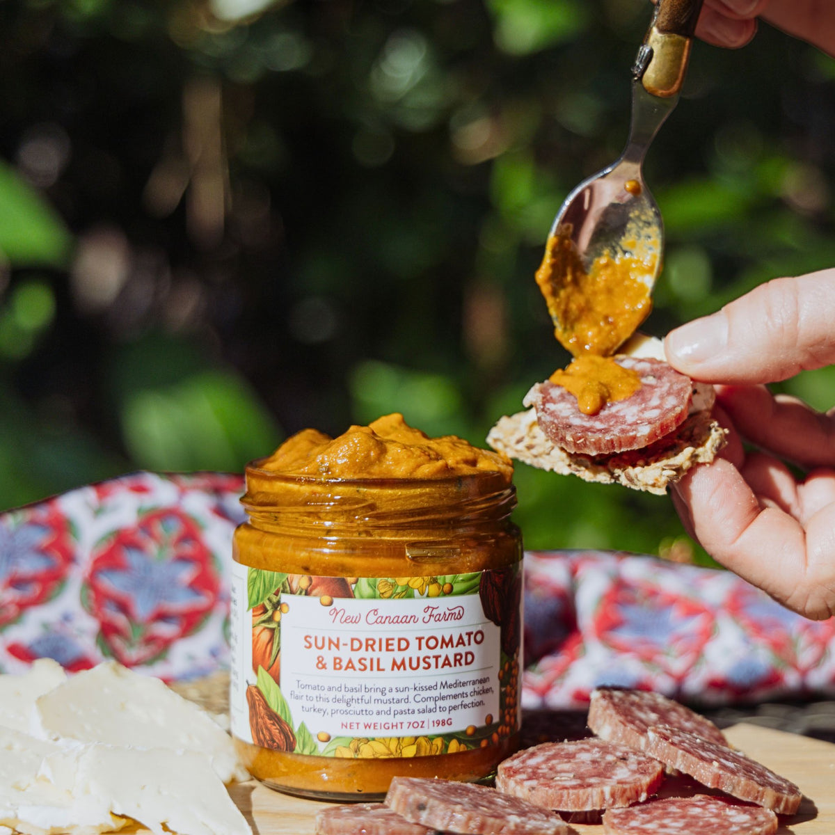 Sun-dried tomato and basil mustard being spread on a cracker with salami.