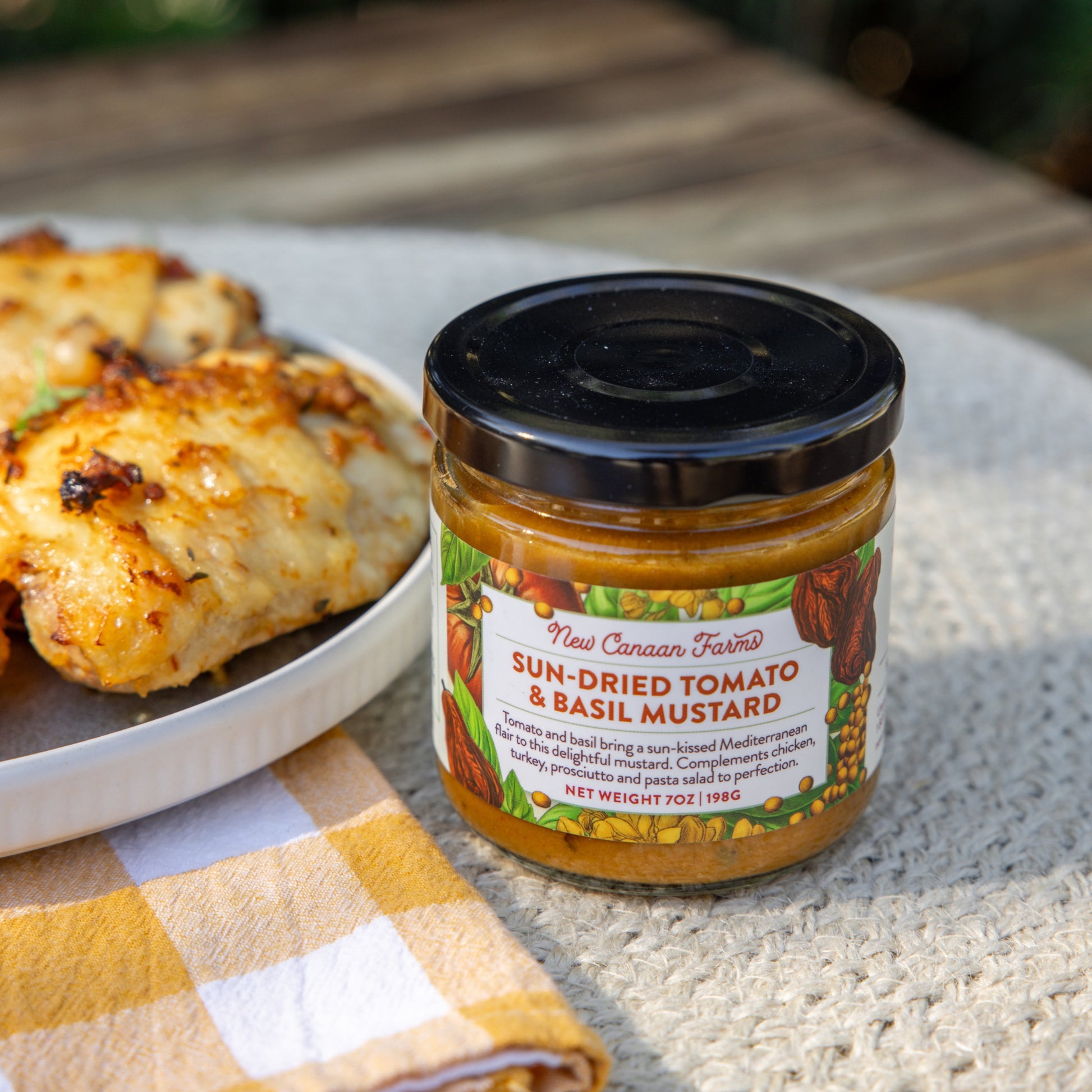 Jar of New Canaan Farms sun-dried tomato and basil mustard on a table with food in the background