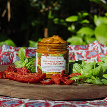 Jar of sun-dried tomato and basil mustard on a wooden board with fresh ingredients outdoors.