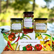 Three jars of New Canaan Farms spreads arranged on top of a vegetable-inspired gift box outdoors, with a blurred green garden background. The jars include Green Olive Tapenade, Lemon Fig Jam, and Goat Cheese & Herbs de Provence Mustard.