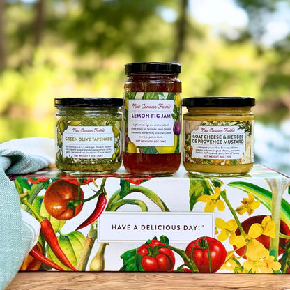 Three jars of New Canaan Farms spreads arranged on top of a vegetable-inspired gift box outdoors, with a blurred green garden background. The jars include Green Olive Tapenade, Lemon Fig Jam, and Goat Cheese & Herbs de Provence Mustard.