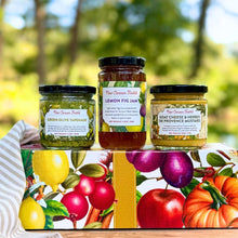 Three jars of New Canaan Farms spreads arranged on top of a seasonal fruits-inspired gift box outdoors, with a blurred green garden background. The jars include Green Olive Tapenade, Lemon Fig Jam, and Goat Cheese & Herbs de Provence Mustard.