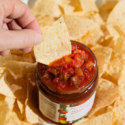 Person dipping a tortilla chip into a jar of salsa with more chips in the background.
