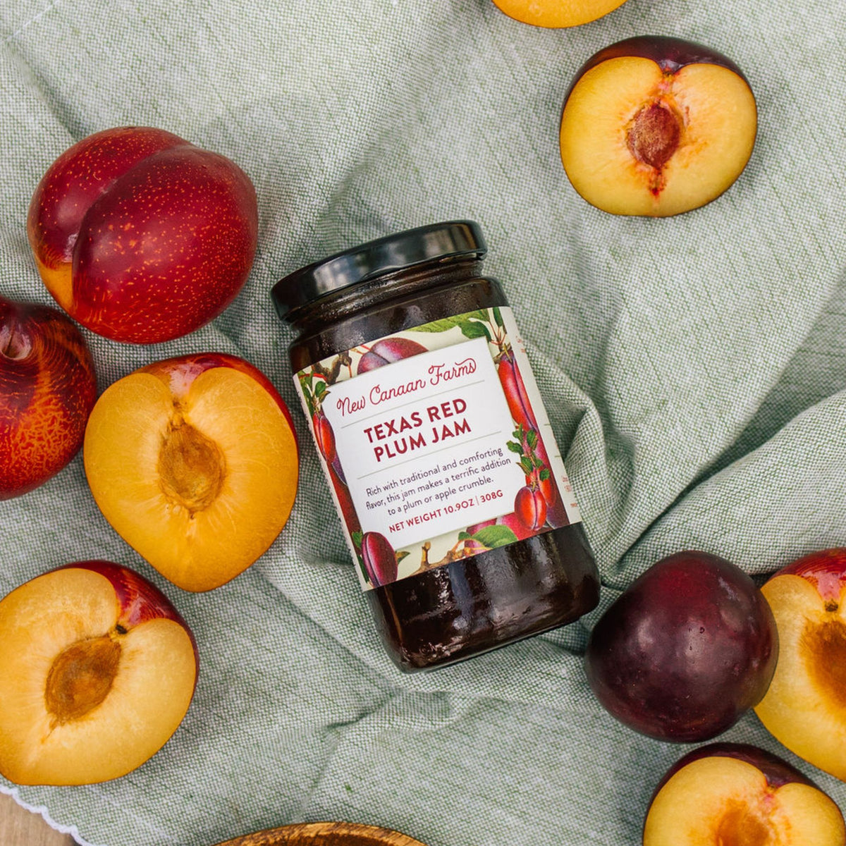 New Canaan Farms Texas Red Plum Jam jar on a green linen cloth among whole and halved plums