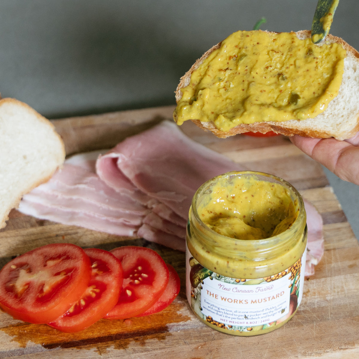 New Canaan Farms The Works Mustard spread on bread with a jar of mustard and ingredients on a wooden board