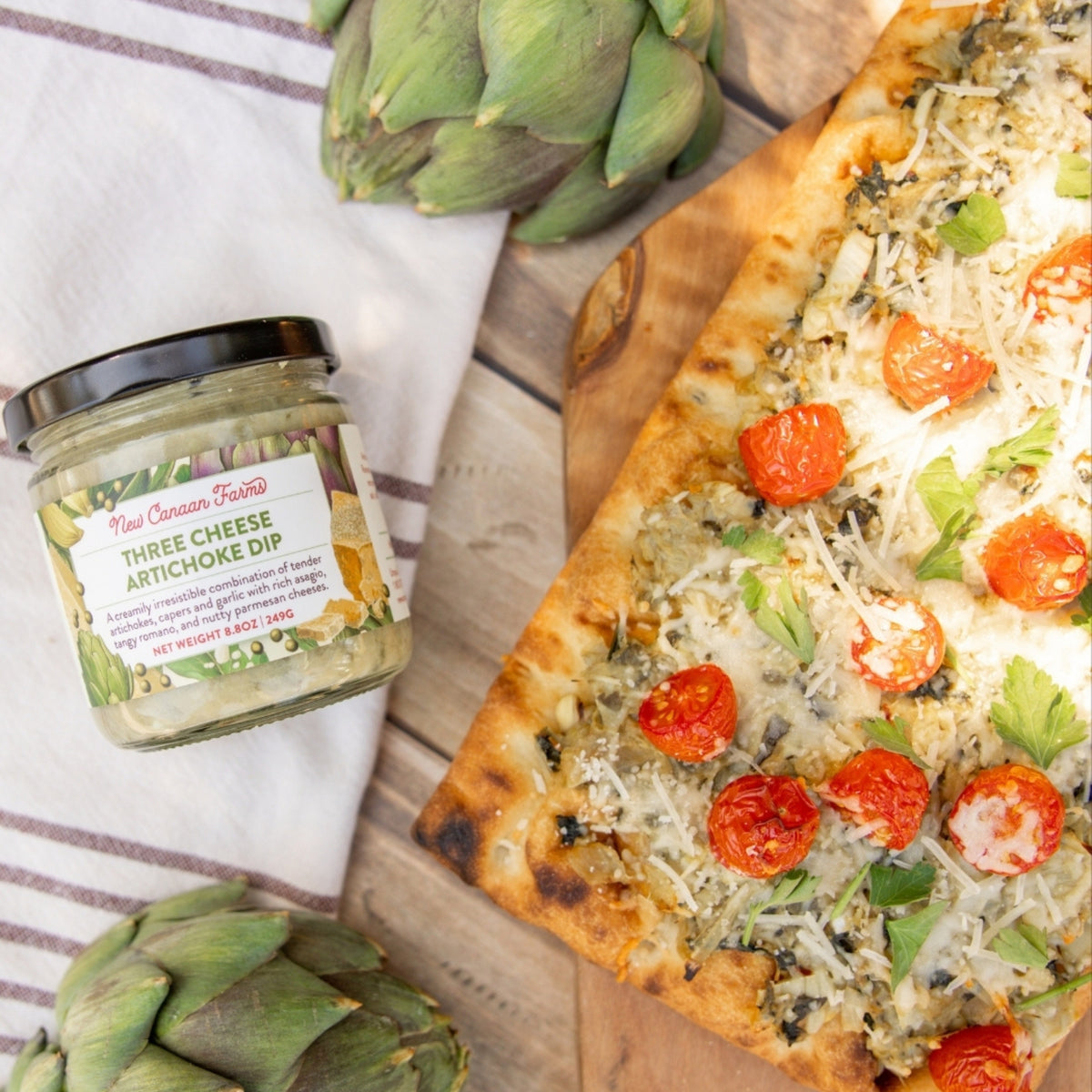 Three Cheese Artichoke Dip jar next to a pizza with artichokes on a wooden board