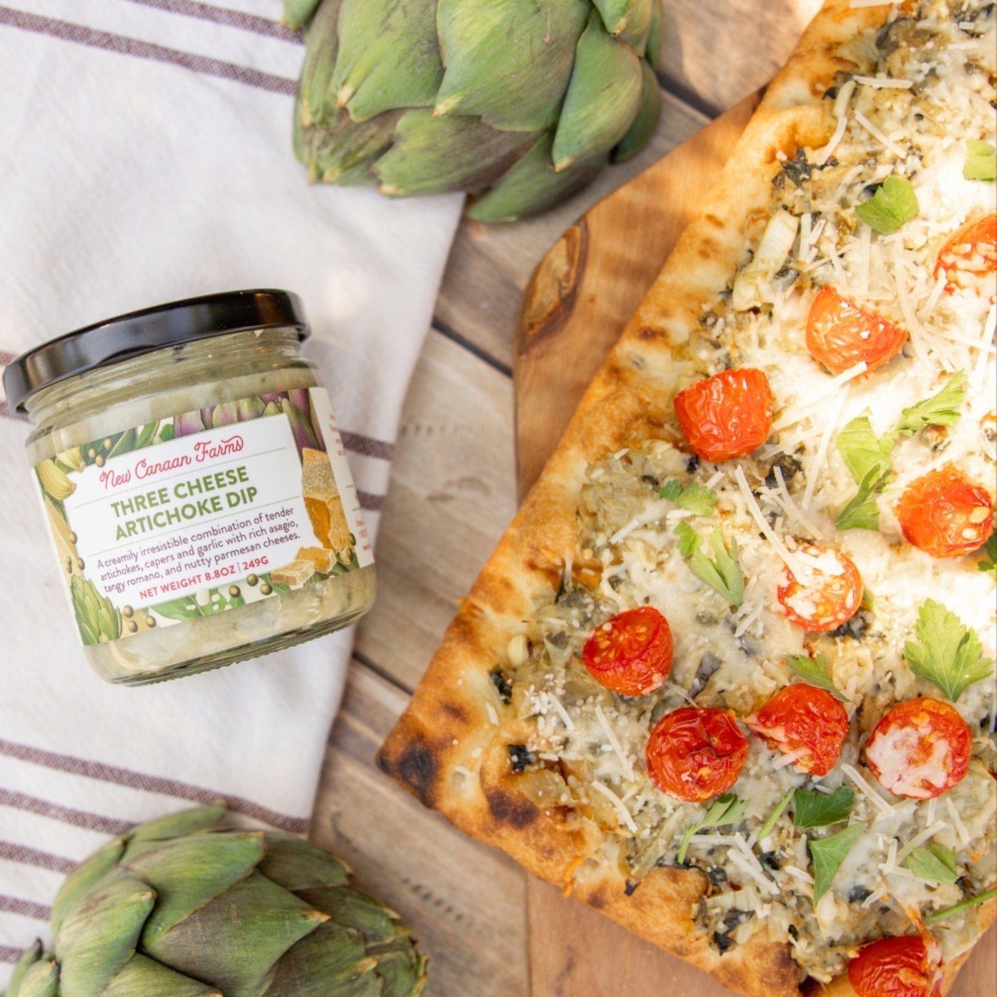 Three Cheese Artichoke Dip jar next to a pizza with artichokes on a wooden board