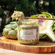 Jar of Three Cheese Artichoke Dip with artichokes on a wooden board