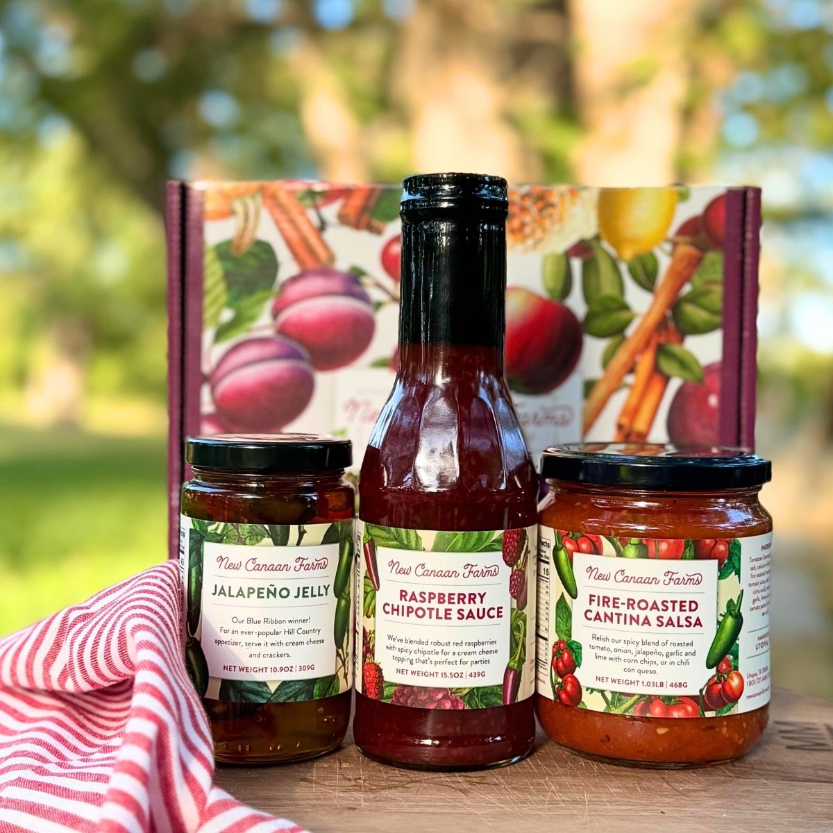 Three jars of New Canaan Farm's most popular products - Jalapeño Jelly, Raspberry Chipotle Sauce and Fire-Roasted Cantina Salsa, in front of a colorful gift box, with a blurred outdoor garden background. A red and white striped cloth is partially visible on the left.