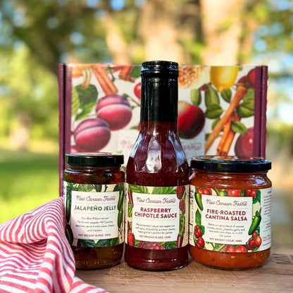 Three jars of New Canaan Farm's most popular products - Jalapeño Jelly, Raspberry Chipotle Sauce and Fire-Roasted Cantina Salsa, in front of a colorful gift box, with a blurred outdoor garden background. A red and white striped cloth is partially visible on the left.