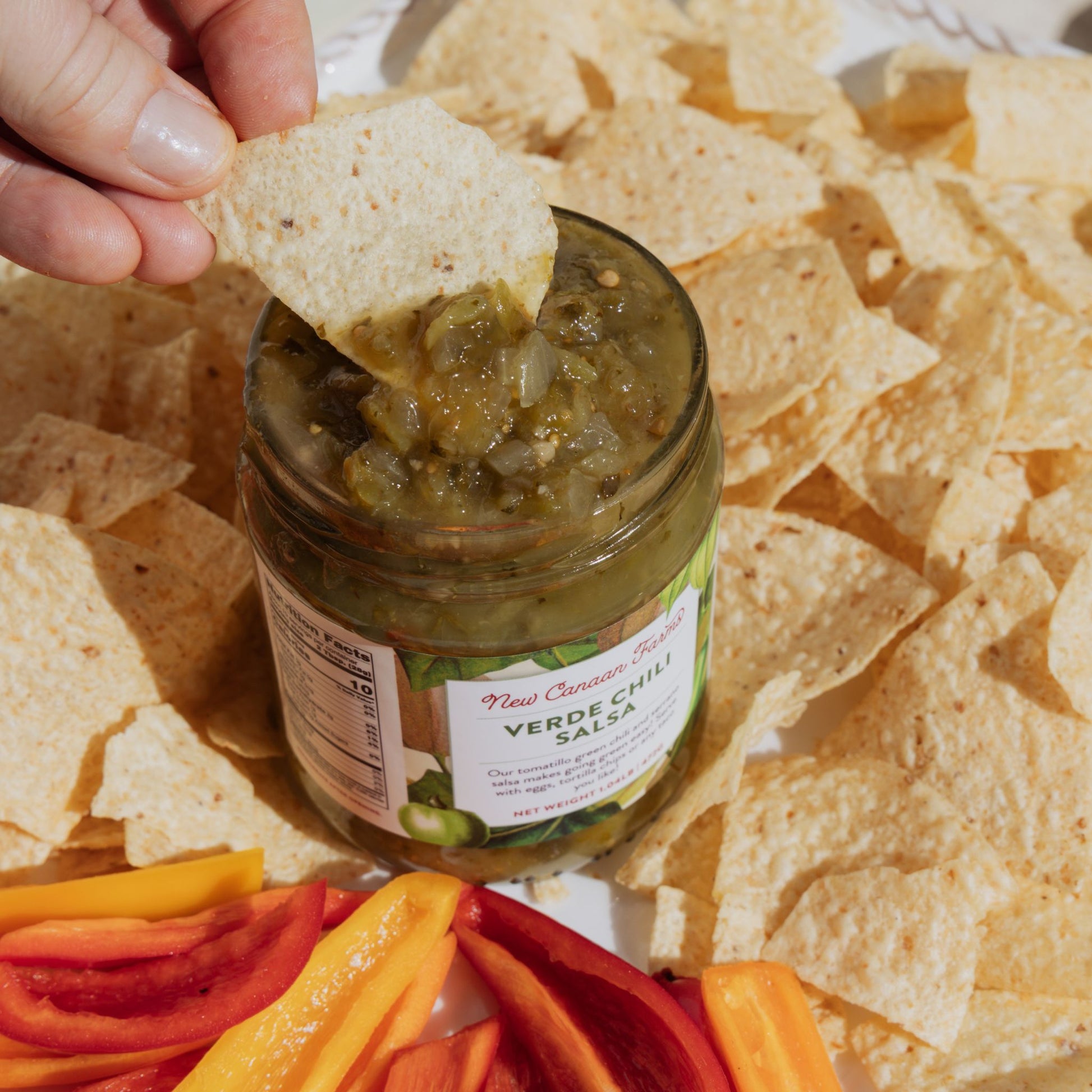 Jar of verde chili salsa with a tortilla chip being dipped into it, surrounded by tortilla chips and sliced bell peppers.
