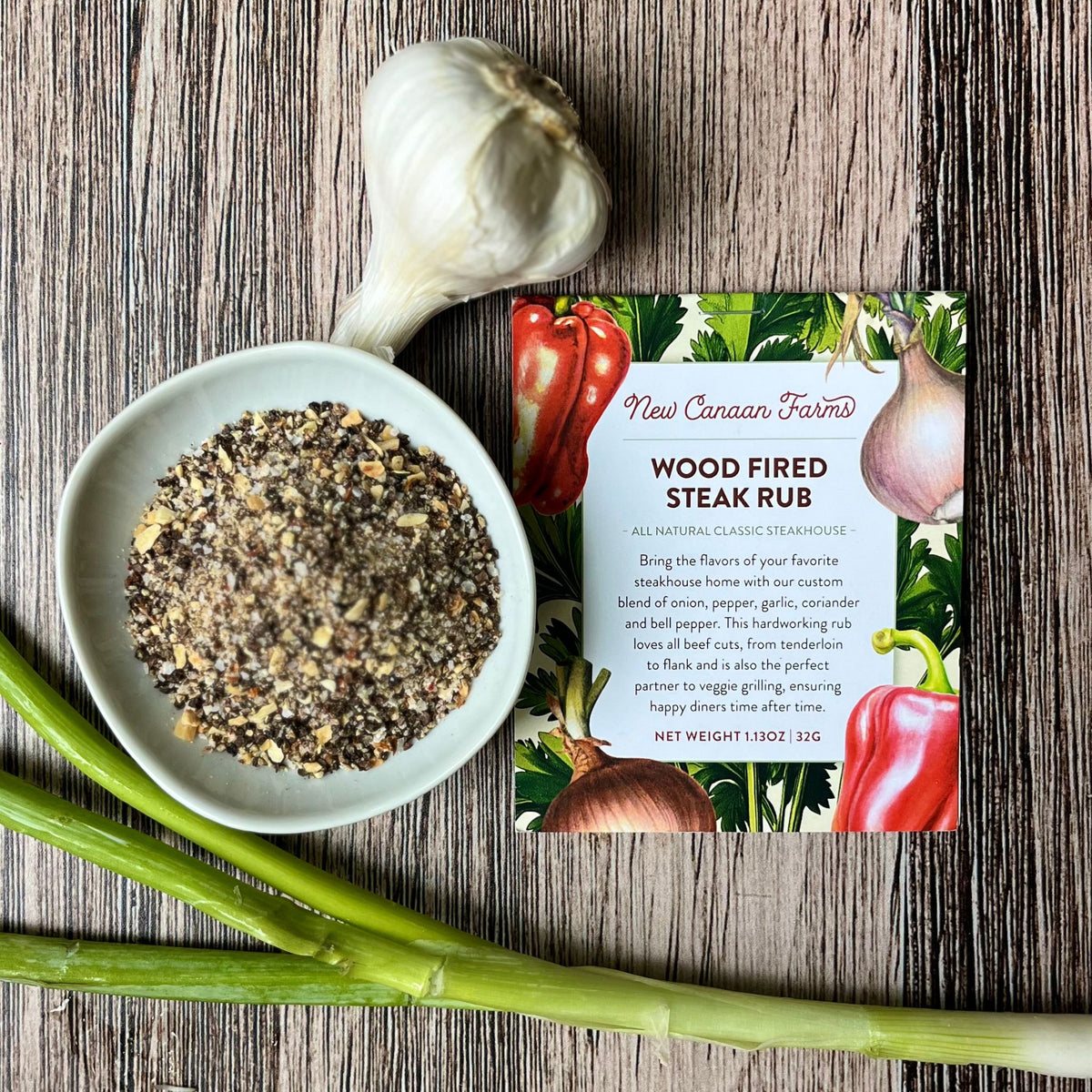 Wood-Fired Steak Rub packet beside a bowl of the coarse rub, a garlic bulb and green onions on a wood table