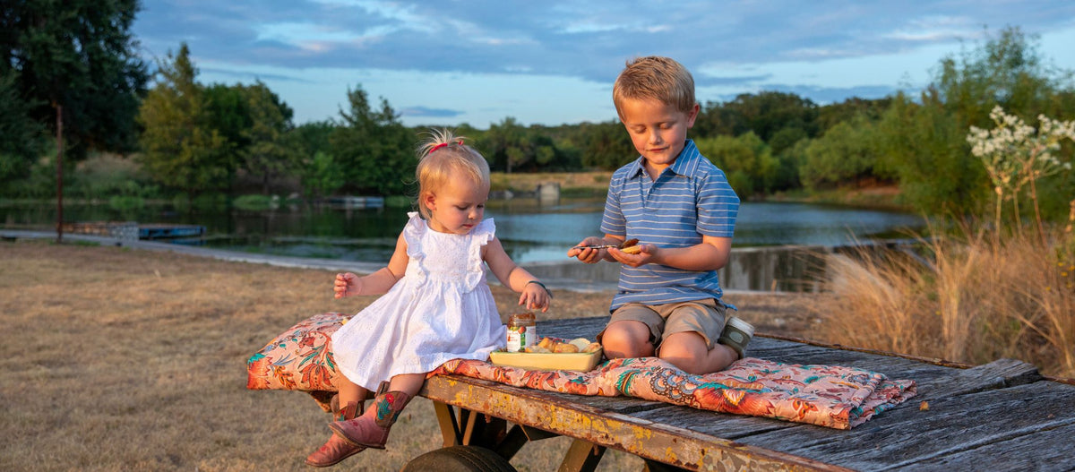 A toddler and a young boy sharing a snack on a quilt-covered picnic table beside a pond