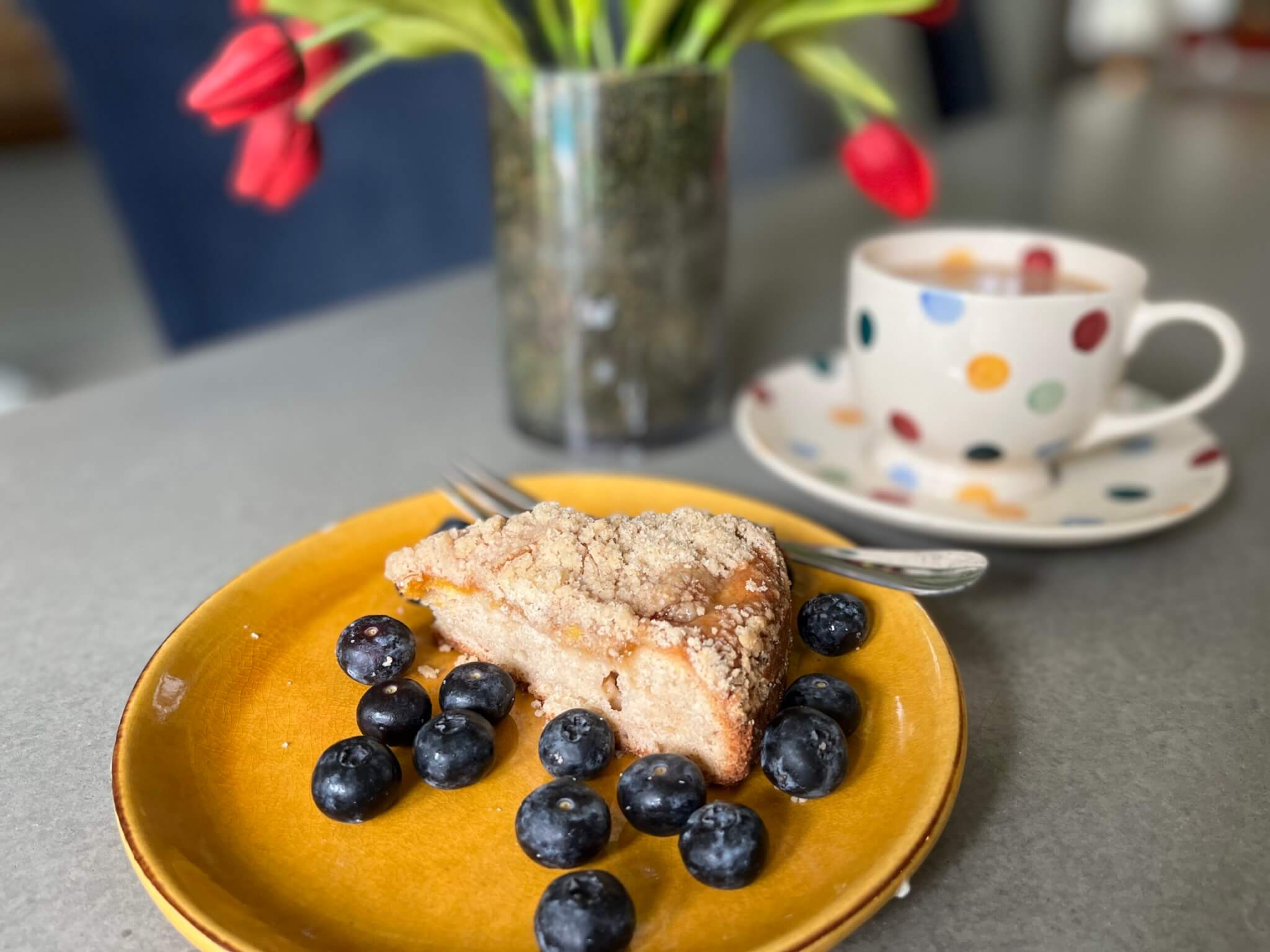 A slice of New Canaan Farms Fredericksburg Peach Jam coffee cake, served with blueberries and a cup of hot tea