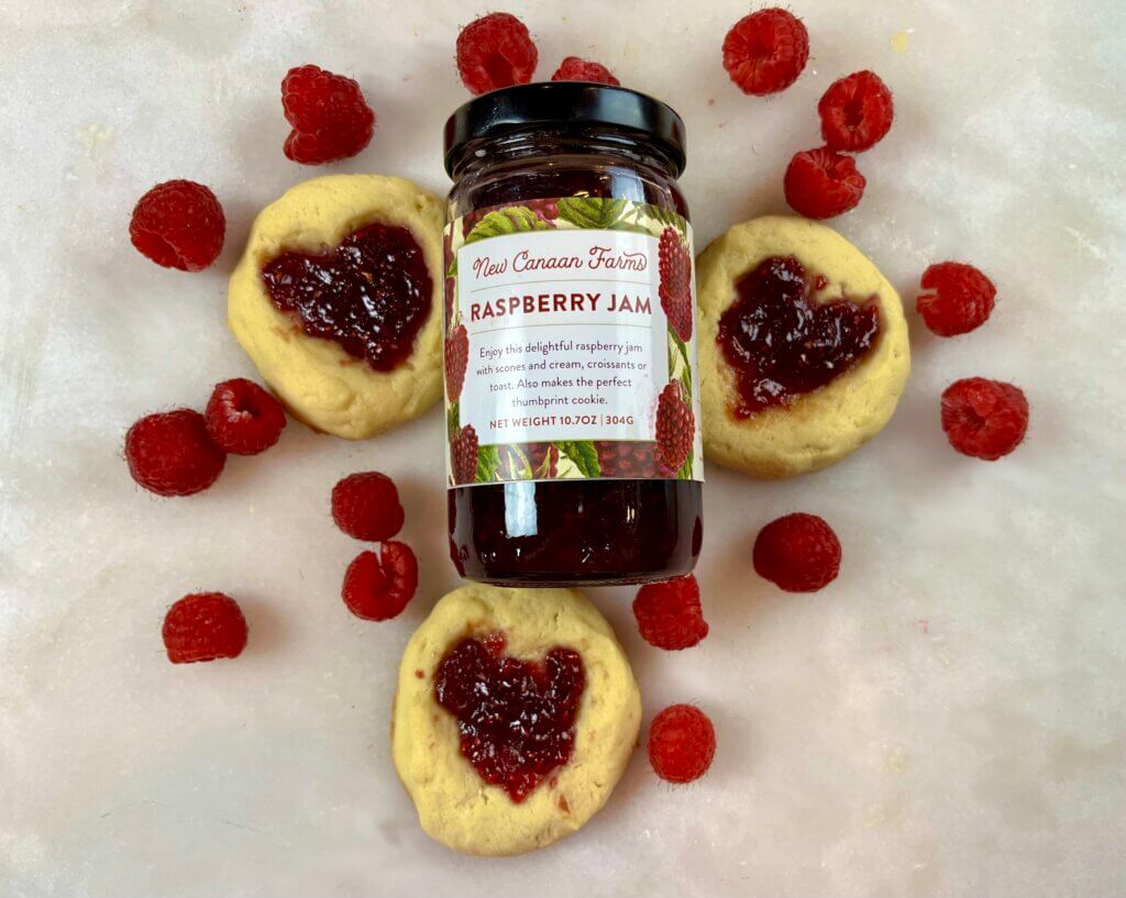 A jar of New Canaan Farms Raspberry Jam surrounded with jam-filled Valentine cookies and fresh raspberries
