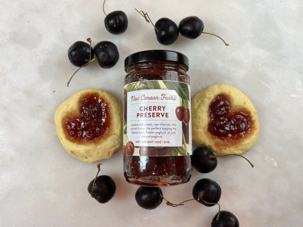 A jar of New Canaan Farms Cherry Preserves surrounded by jam-filled Valentine cookies and fresh cherries