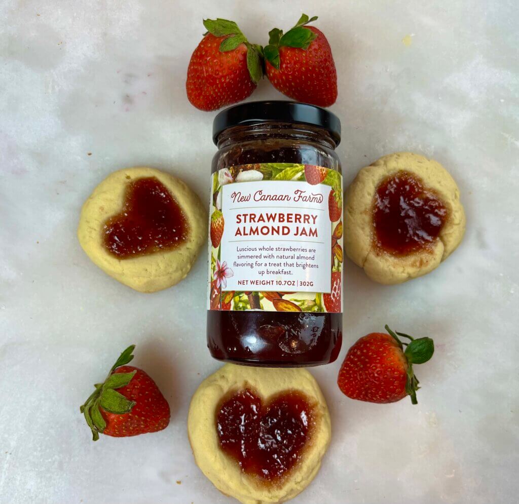 A jar of New Canaan Farms Strawberry Alomd Jam surrounded by Thumbprint Valentine Cookies and fresh strawberries