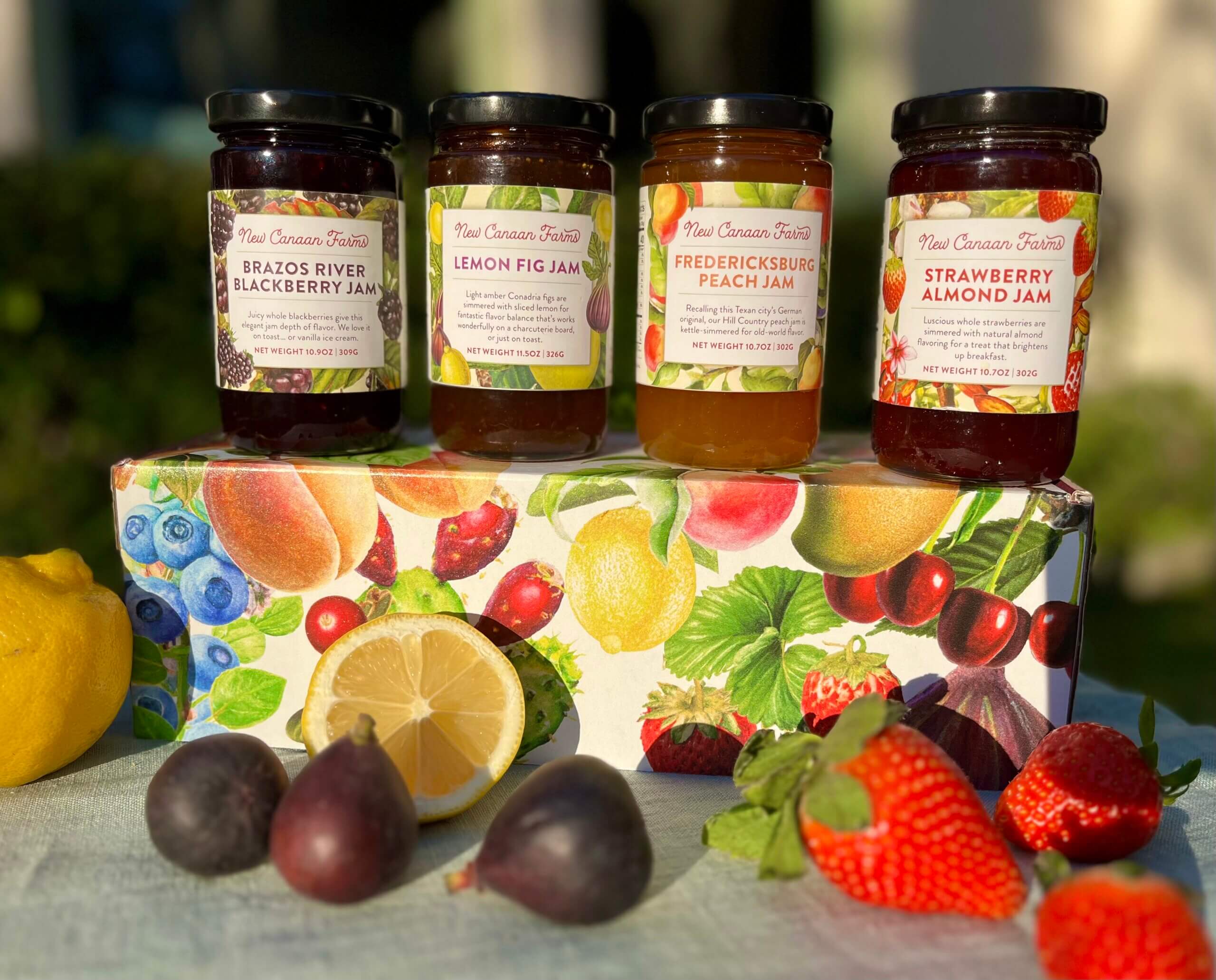 Four New Canaan Farms customer favorite jams on top of a gift box with scattered fruit around them; Blackberry, Lemon Fig, Peach and Strawberry Almond