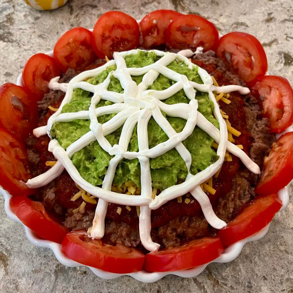 A white pie dish filled with refried beans at the base, covered with New Canaan Farms Cilantro Salsa, then cheddar cheese, then mashed avocado and finally a cobweb of sour cream
